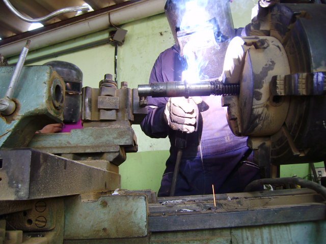 Persona trabajando en un torno mecánico, usando equipo de protección y soldando una pieza metálica.