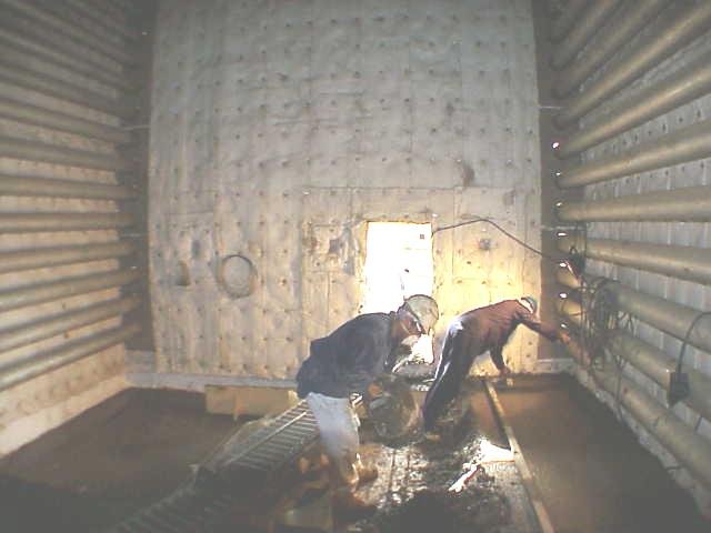 Vista interior personas trabajando en un horno en una construcción, una de ellas extendiendo concreto en el suelo.a y piso de concreto en proceso de construcción, con algunas herramientas y un balde.