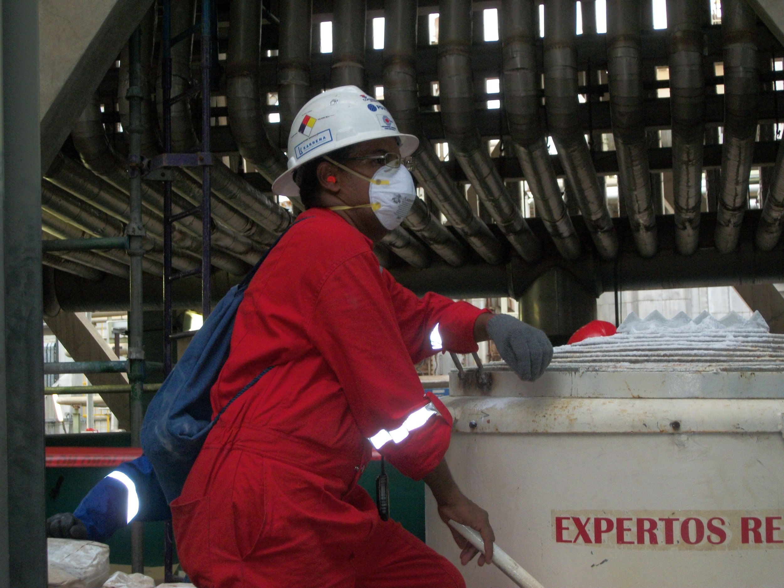 Trabajador usando casco, mascarilla y guantes en una planta industrial, con tubos y maquina mezcladoraen el fondo.