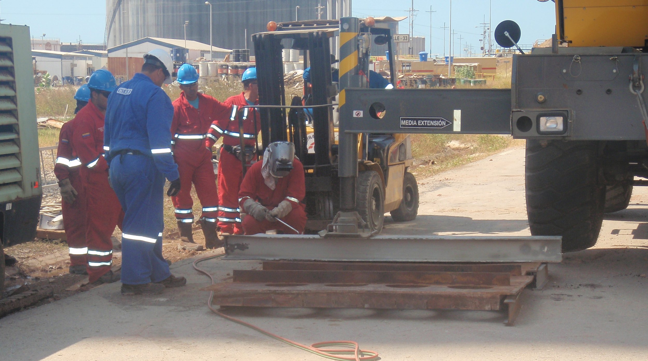 Trabajadores en uniforme de seguridad, algunos con cascos, operan una máquina de construcción y trabajan en una superficie de concreto en un área industrial o de construcción.