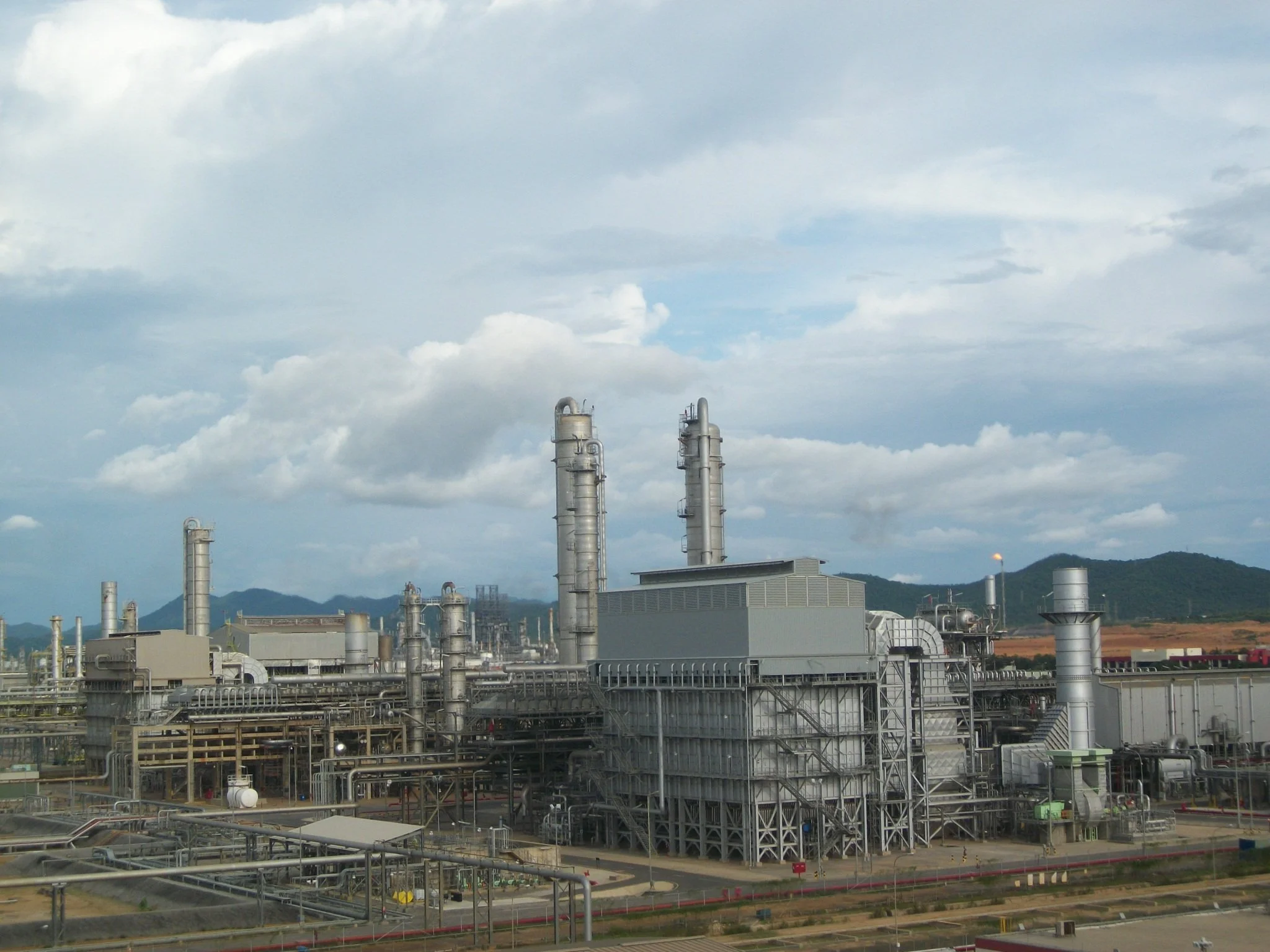 Planta industrial con torres de acero y tuberías en un paisaje montañoso, cielo nublado.