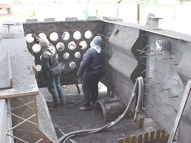 Dos trabajadores en equipo usando casco, gafas y máscara de protección, trabajando en una Estructura de acero con anclajes refractarios en un sitio de construcción.
