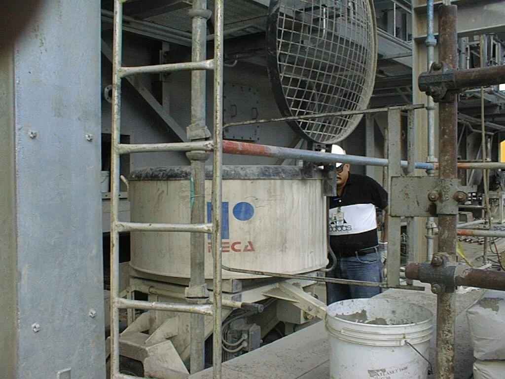 Una máquina de mezcla de concreto en un sitio de construcción rodeada de andamios y tuberías, con una persona parcialmente visible detrás.