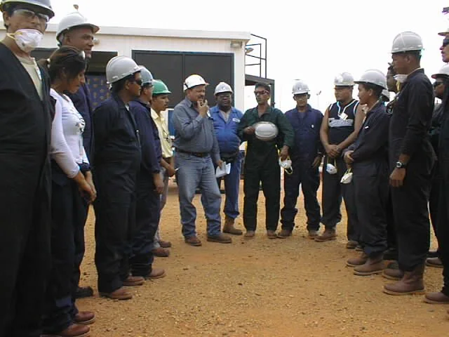 Grupo de personas con casco y mascarillas en una visita de inspección en una obra de construcción o sitio industrial, formando un círculo y escuchando a un guía.