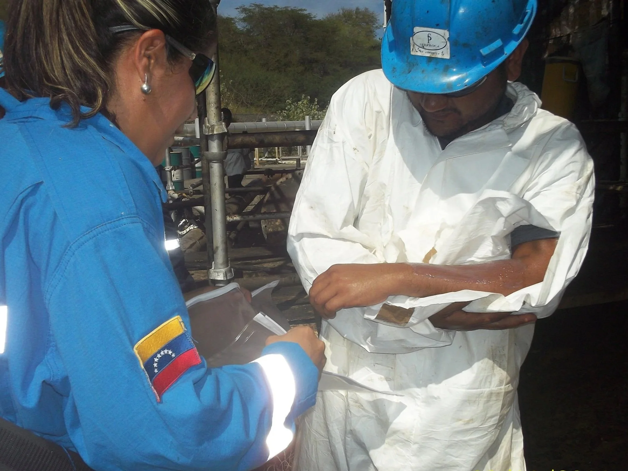 Mujer con uniforme azul y parche de Venezuela revisando una muestra en presencia de un hombre con traje blanco y casco azul, en un entorno industrial o científico.