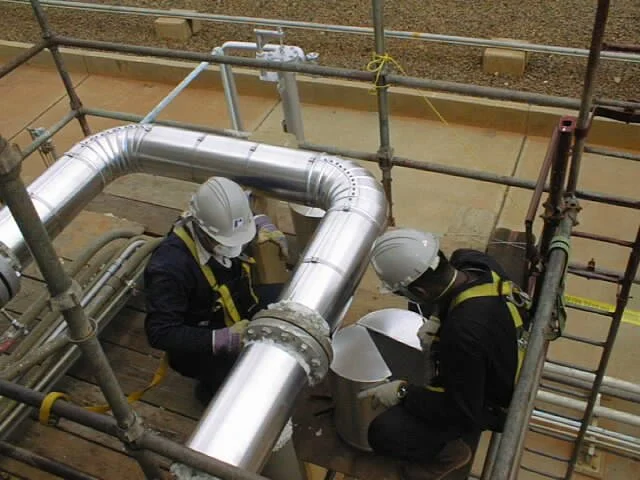 Dos trabajadores con casco y arnés instalando tuberías de metal en una plataforma de construcción.