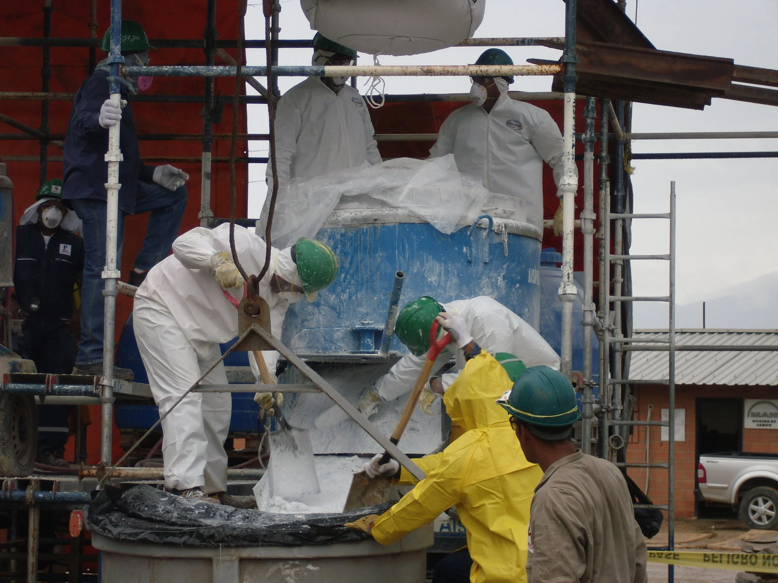 Trabajadores en un sitio de construcción manipulan concreto con equipo de protección, incluyendo cascos y guantes, en un escenario con estructuras de andamios y edificios en el fondo.