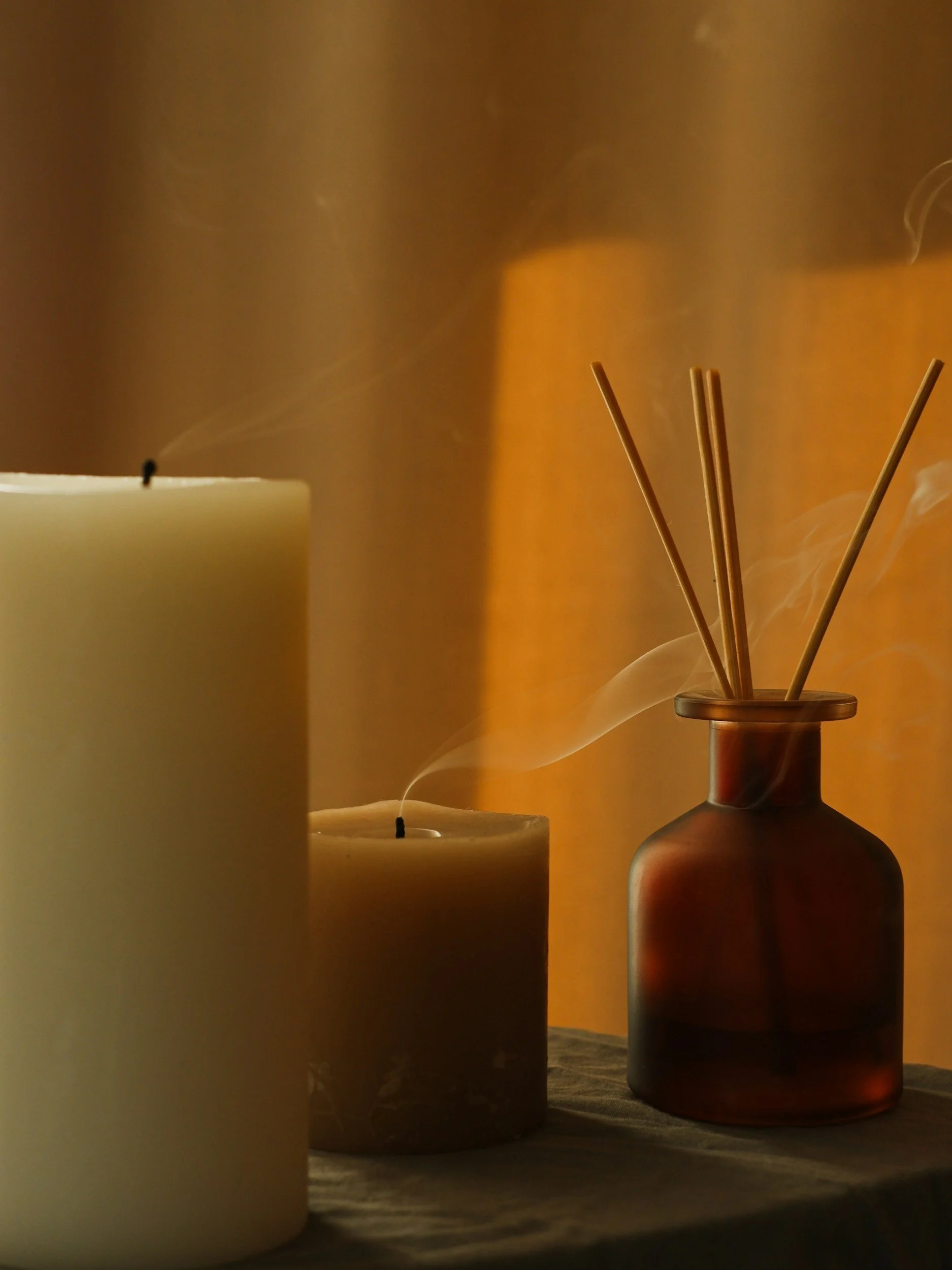 Three candles and a reed diffuser on a wooden surface with warm lighting and visible smoke.