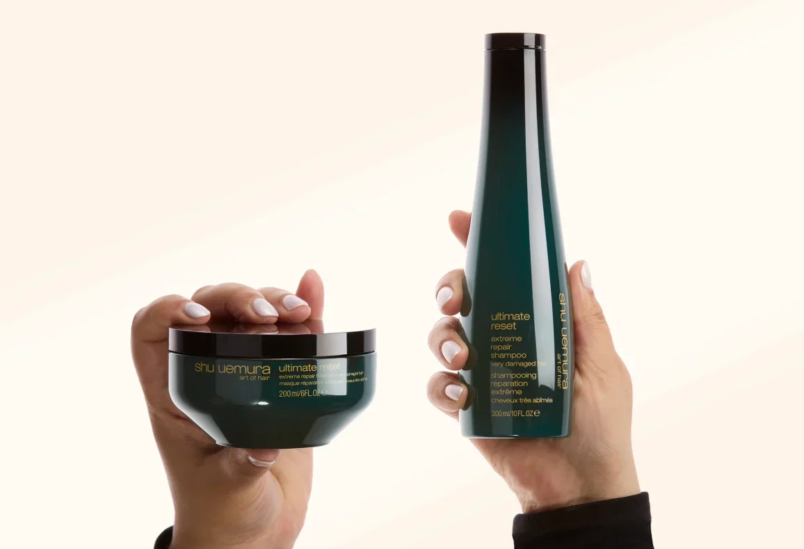 Two hands holding bottles of Shu Uemura Ultimate Reset hair care products, including a jar and a tall bottle, against a light background.