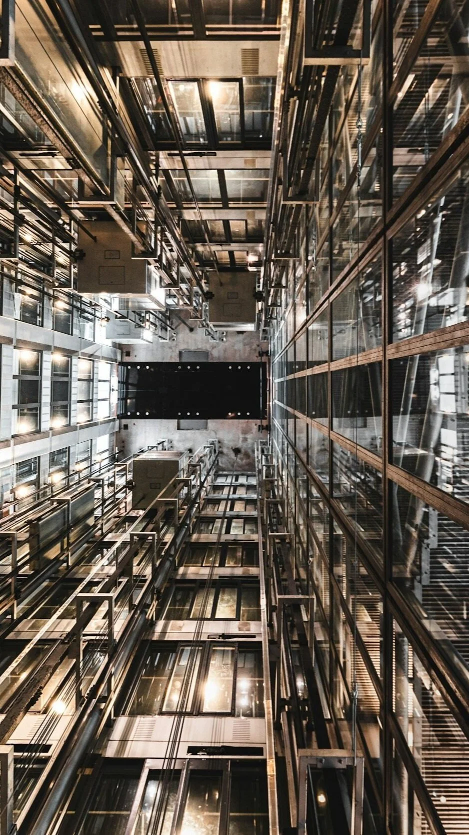 View looking down into a modern, glass-enclosed elevator shaft with metal and glass architecture, lights, and a black ceiling panel at the bottom.