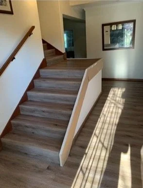 Indoor staircase with wooden steps and white risers, next to a window casting sunlight on the floor, with a framed picture hanging on the wall.