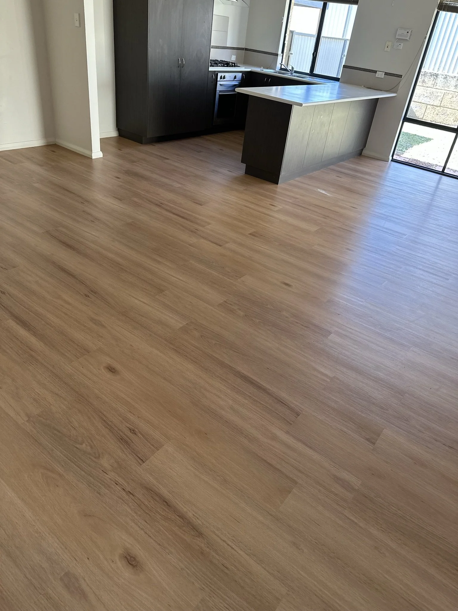 Empty room with wooden flooring and a kitchen in the background, featuring dark cabinets, a stove, sink, and a kitchen island. Large windows let in natural light.