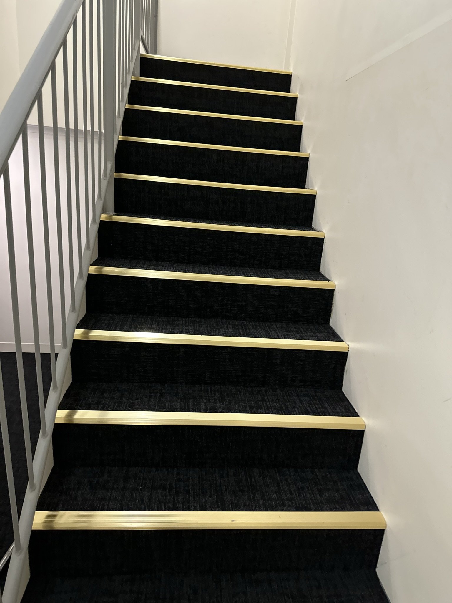 Indoor staircase with black carpeted steps and gold-colored metal trim, white walls, and a white railing on the left side.