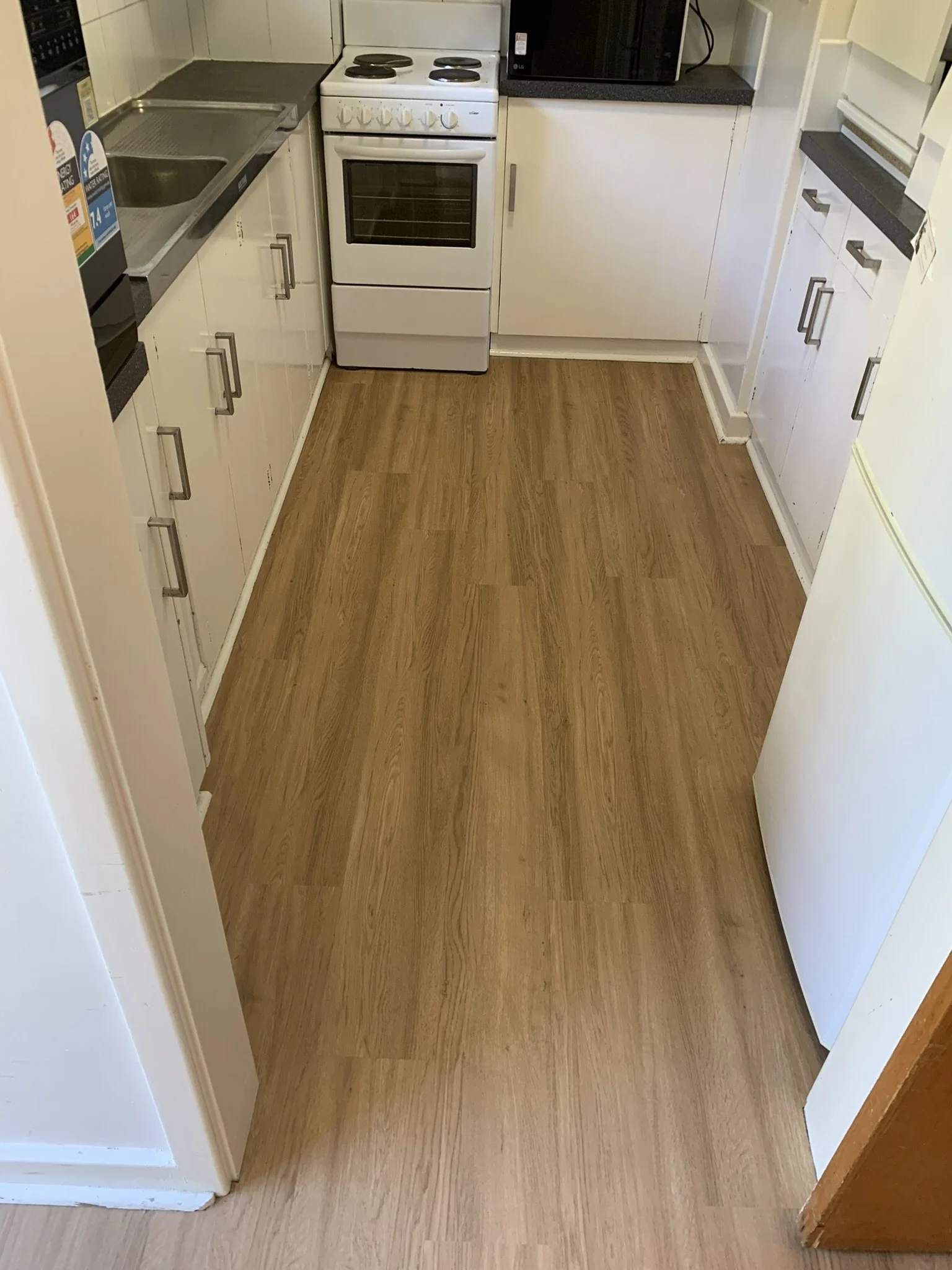 Empty kitchen with wooden floor, white cabinets, a sink, a stove, microwave, and refrigerator.