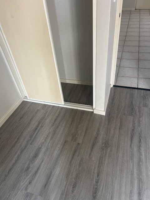 A room with gray wood-like flooring, a closet with a sliding door, and an adjacent kitchen area with white tiles.