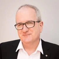 A middle-aged man with gray hair and glasses wearing a black blazer and white shirt, smiling against a plain background.