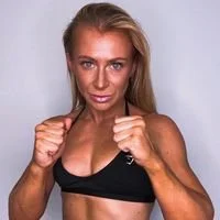 A woman with blonde hair in a fighting stance, wearing a black sports bra, looking at the camera, against a light gray background.