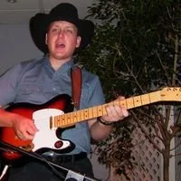 Person wearing a cowboy hat playing an electric guitar outdoors at night.