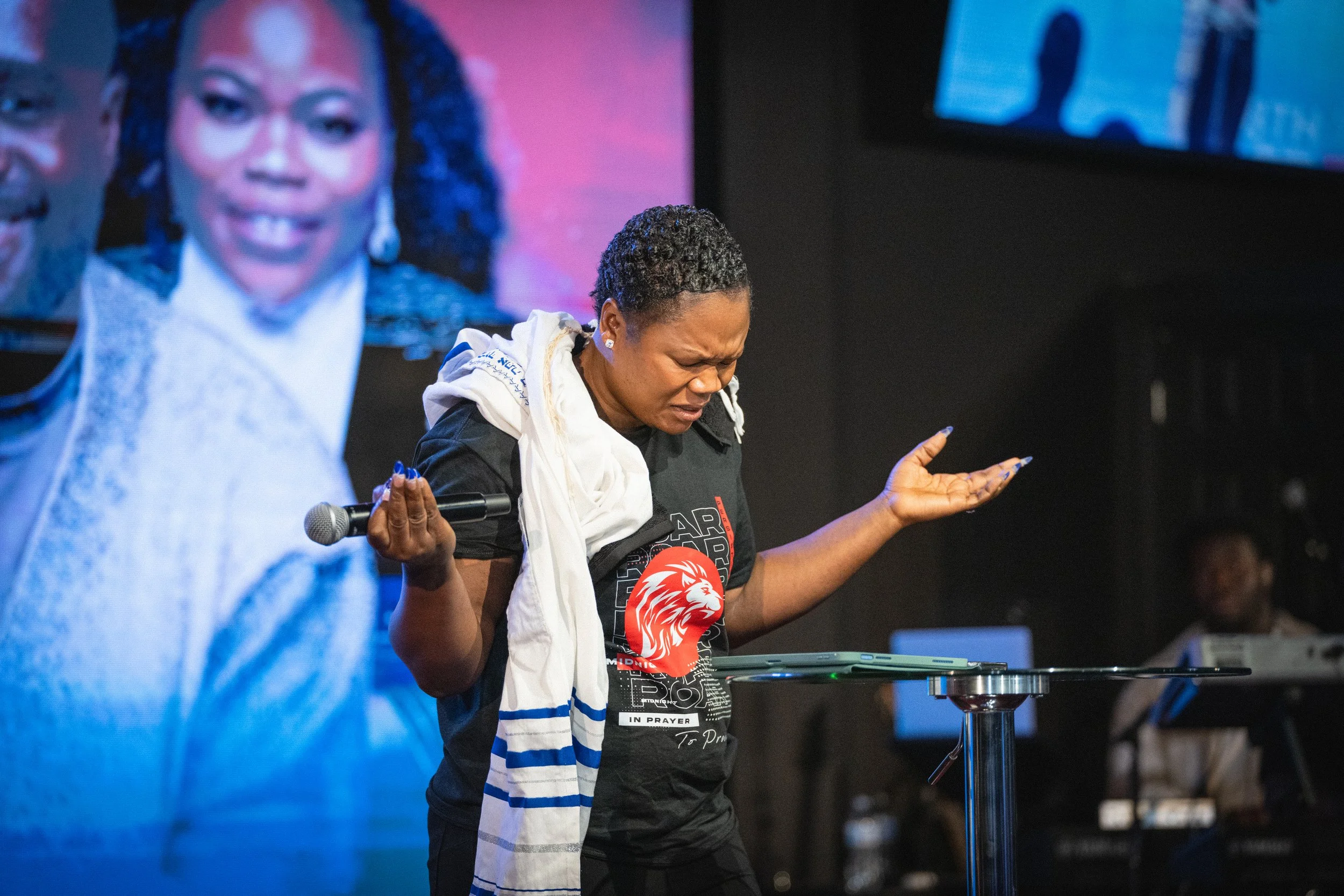 Woman with short curly hair, wearing a black T-shirt with a lion emblem, holding a microphone in her right hand, standing behind a podium, with her eyes closed and a distressed expression, on a stage with large screens showing her face in the background.