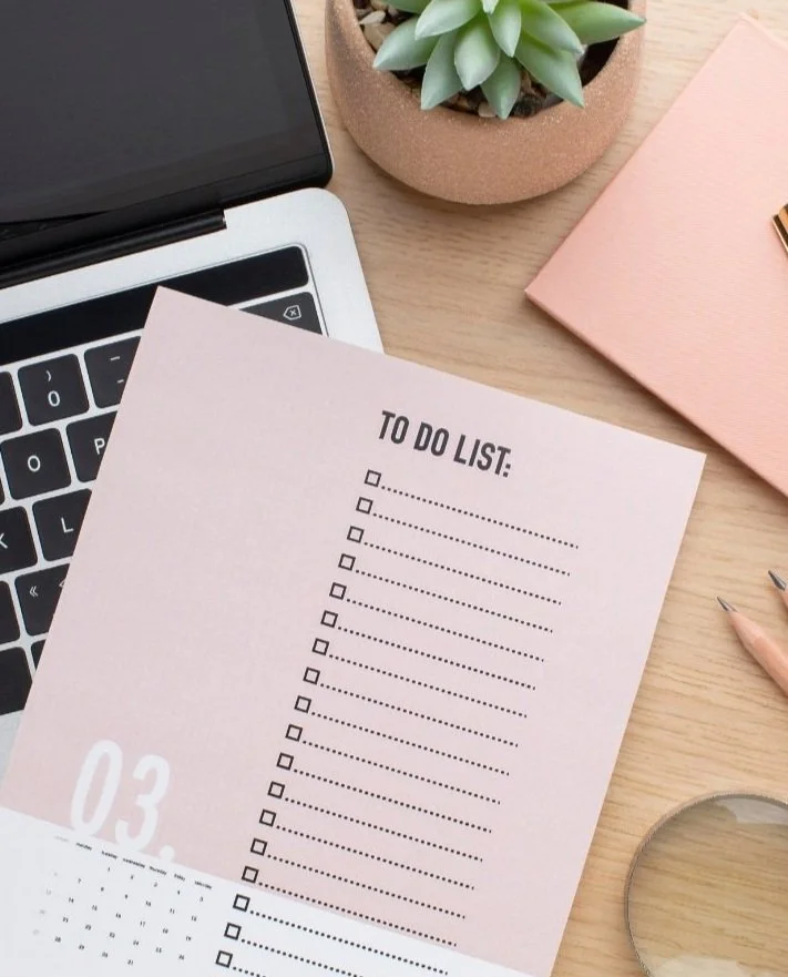Workspace with laptop, to-do list pad, pencils, magnifying glass, succulent plant in pot, and pink notebook on wooden desk.