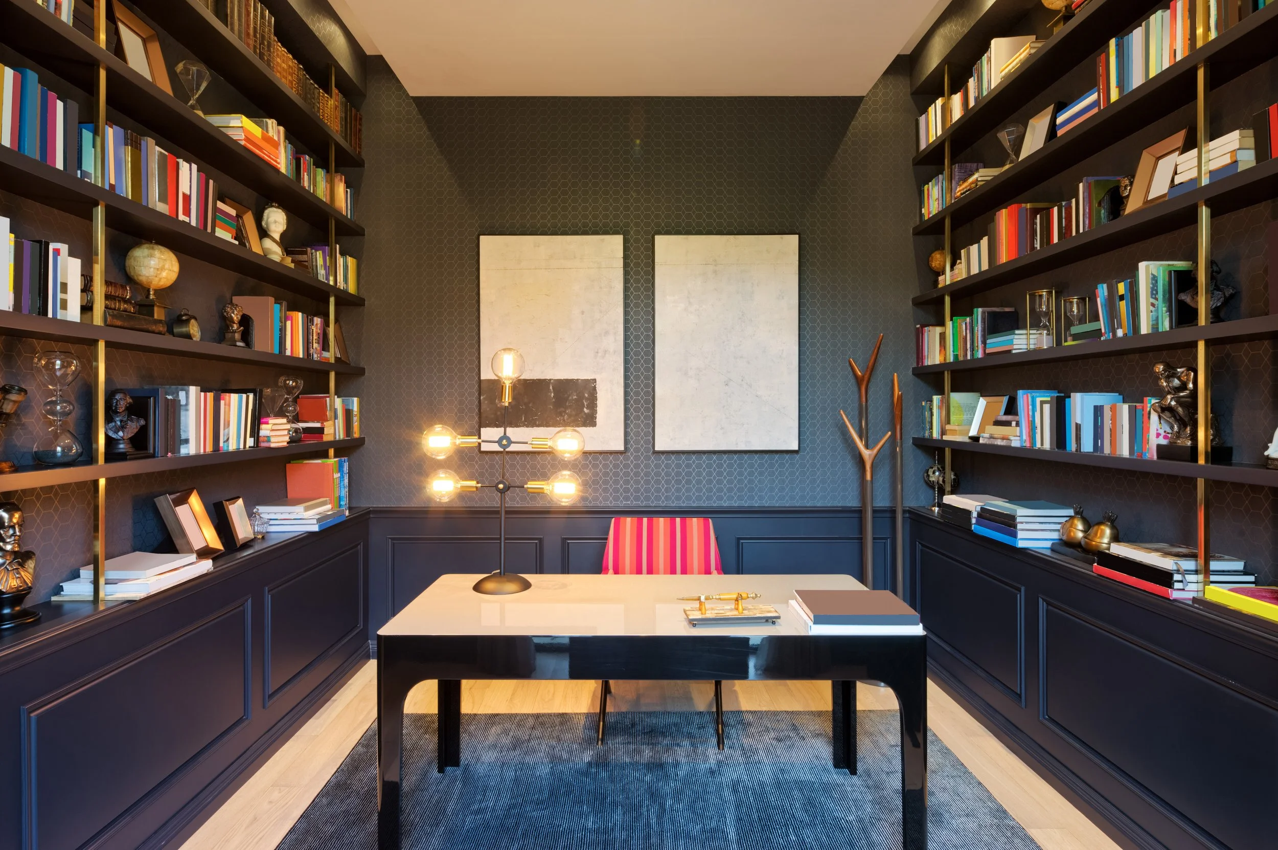 A modern office with dark patterned walls, two large bookshelves filled with colorful books and decorative items frame a white desk with a black border. Behind the desk is a red and pink striped chair, and on the wall are two abstract art pieces. The room has a warm lighting fixture with exposed bulbs and a tall wooden coat rack to the right.
