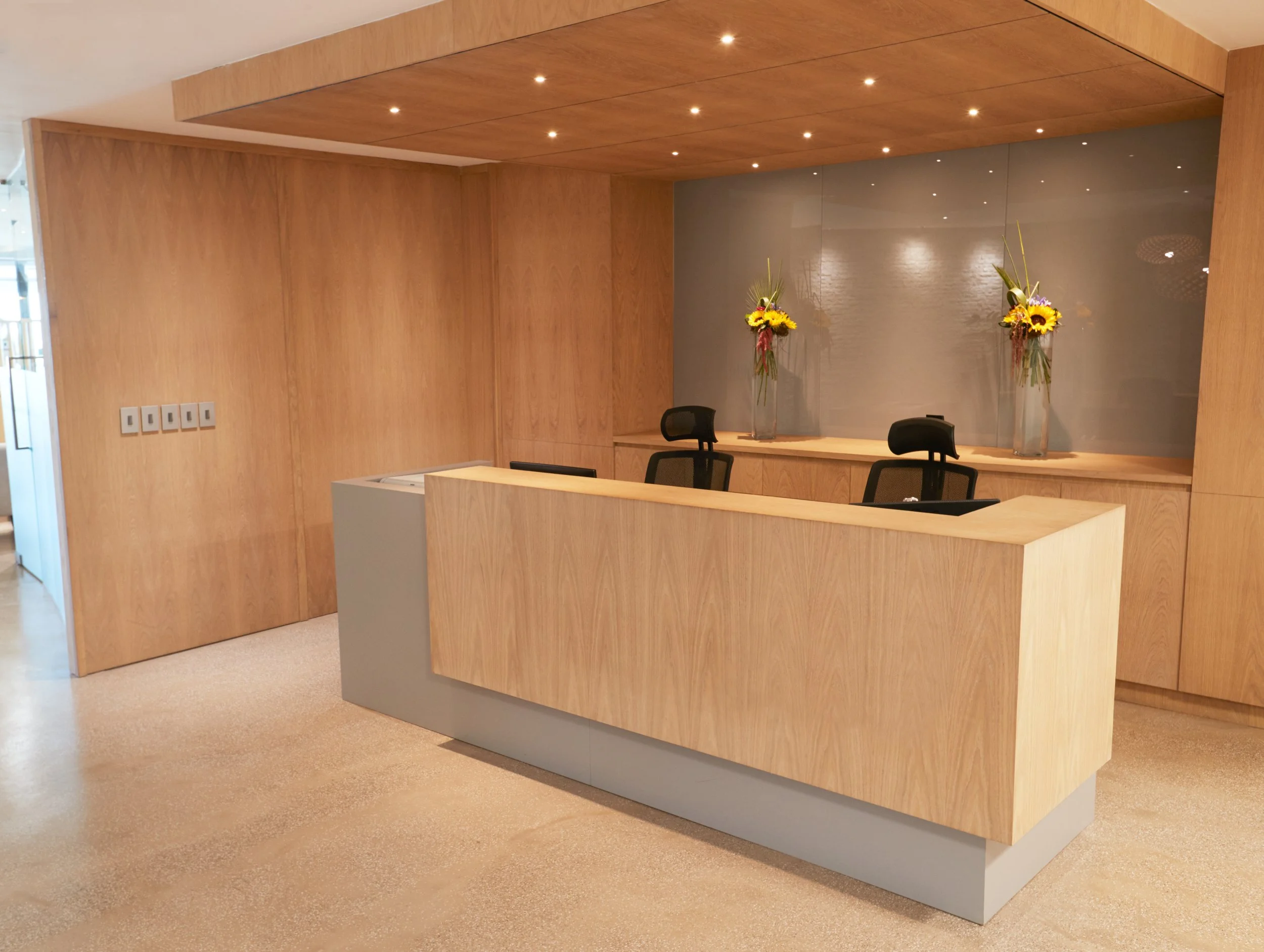 Minimalist reception area with a wooden counter, black chairs, and tall glass vases with sunflowers and other flowers on a driftwood shelf against a fog-gray wall.