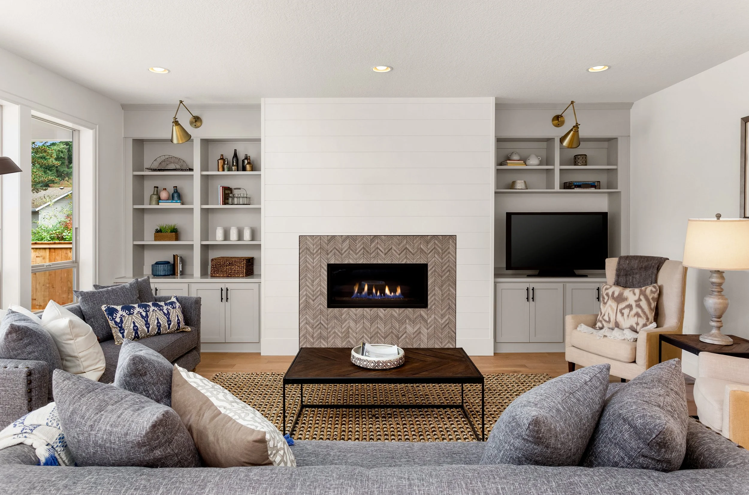 Living room with a modern fireplace, built-in white shelves, gray sectional, beige armchair, a coffee table, and a TV, with windows allowing natural light