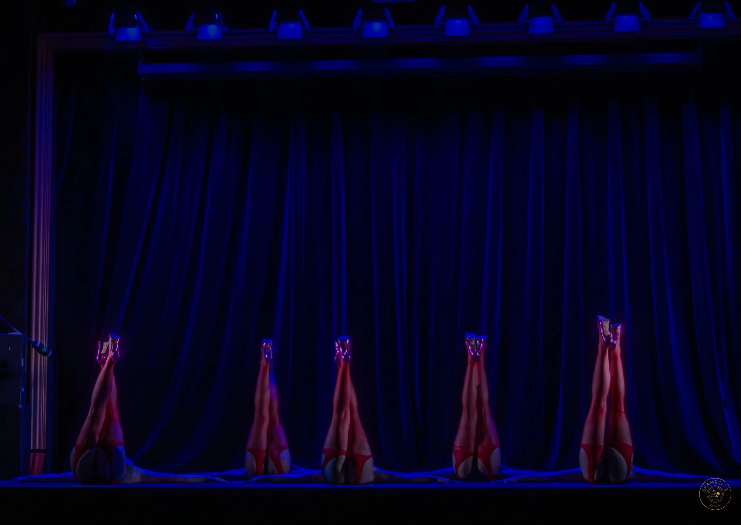 A stage with a dark background and blue curtains, featuring five women lying on their backs with legs raised vertically, wearing red high heels and red outfits.