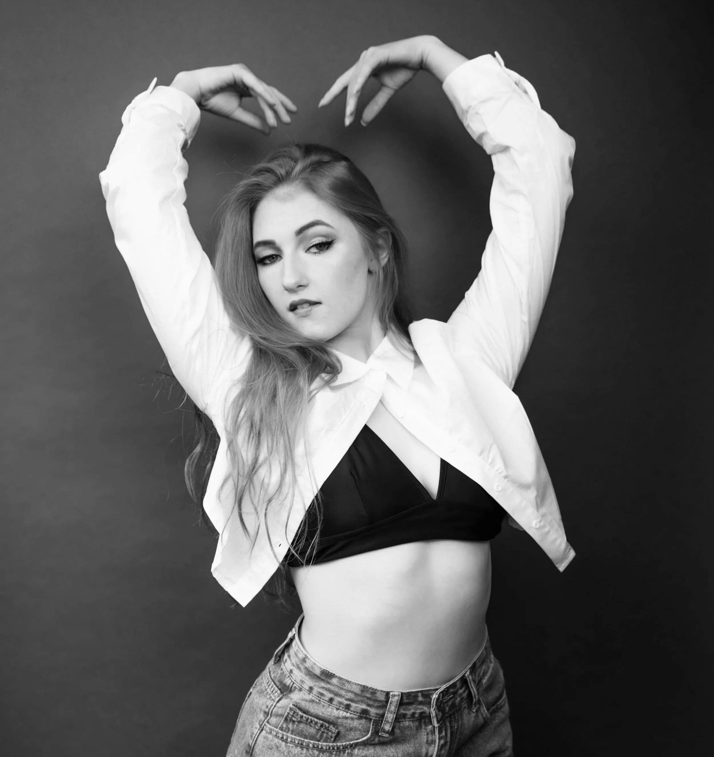 Black and white photo of a young woman with long hair, wearing a white shirt over a black crop top and denim jeans, making a heart shape with her arms raised above her head.