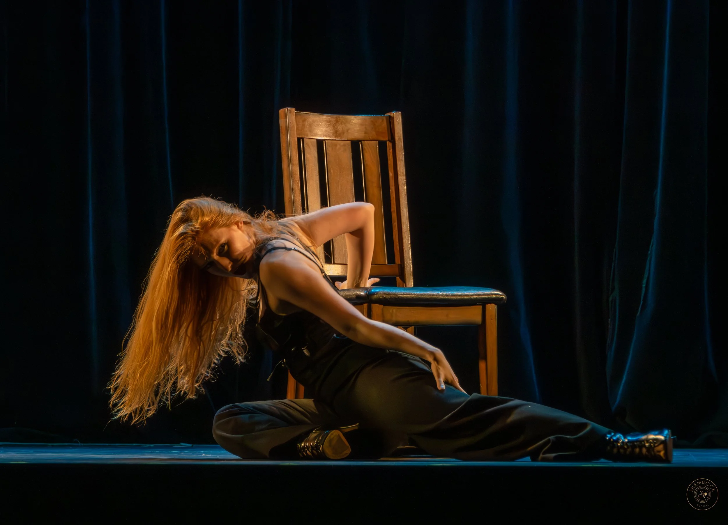A woman with long red hair performs a dance move on a stage, sitting on the floor with one leg bent and the other extended, with a wooden chair behind her. She is wearing a black sleeveless top and black pants, with a dark curtain in the background.