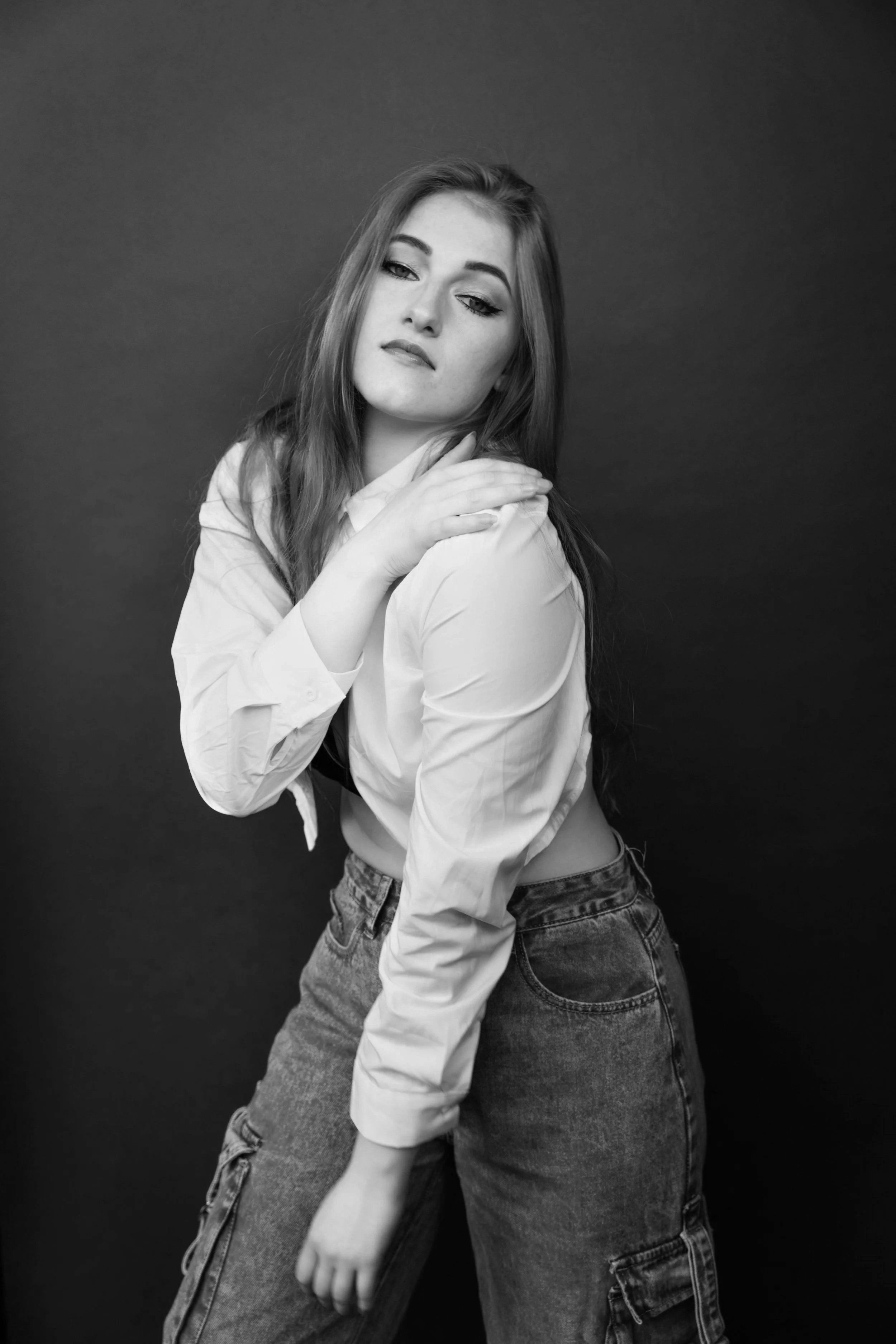 Black and white photo of a young woman with long hair, wearing a white shirt and jeans, posing with her hand on her shoulder against a plain dark background.