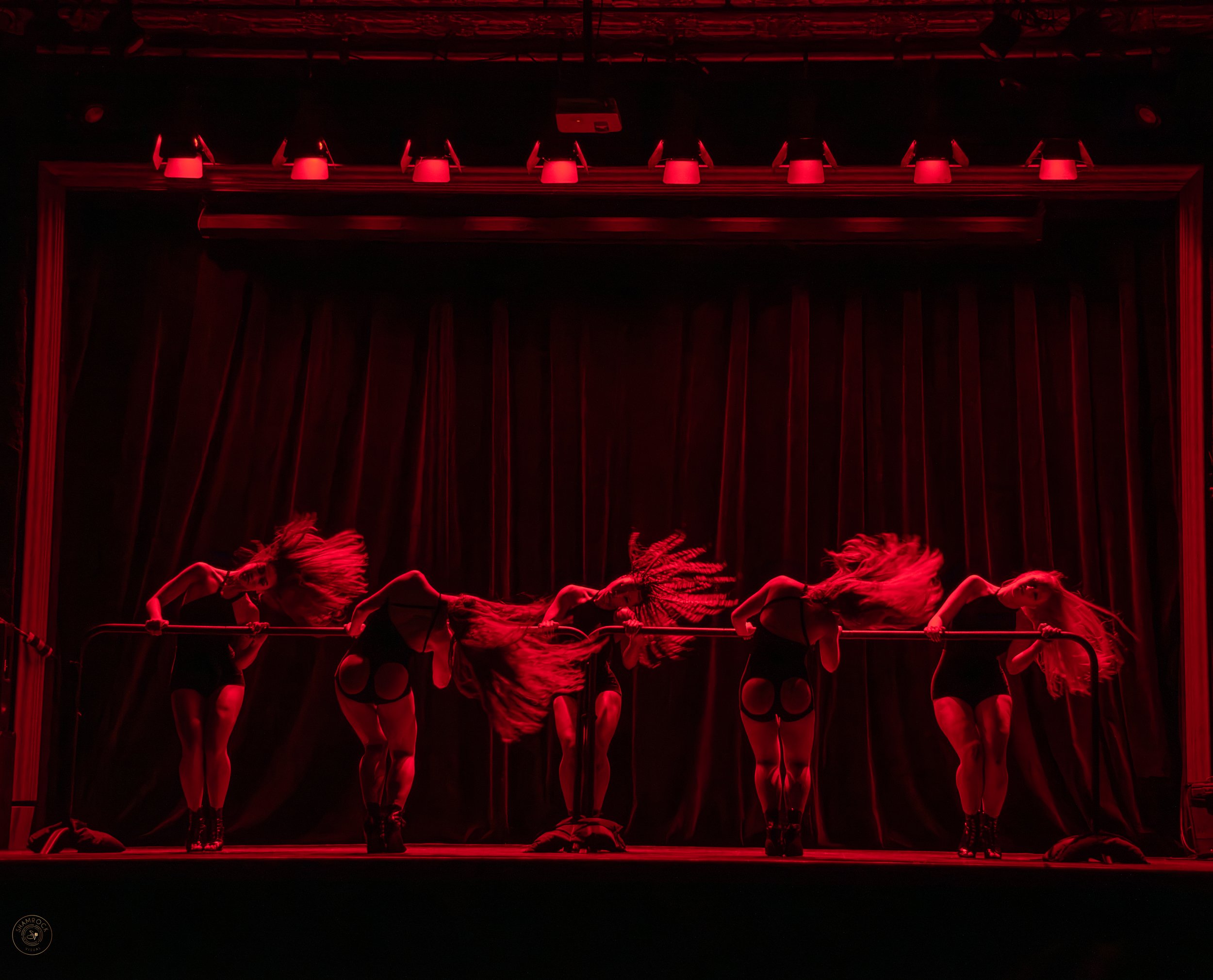 Six female performers with long hair performing on stage under red lighting, bending backward over a horizontal bar, with a dark curtain backdrop.