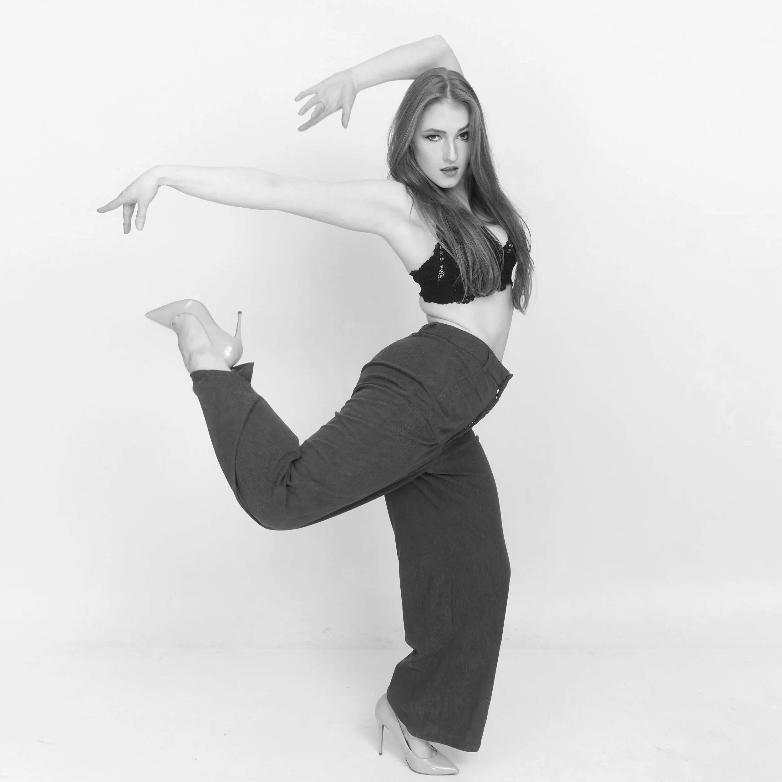 A woman in high heels poses with one leg bent and raised, wearing dark pants and a crop top, against a plain background.