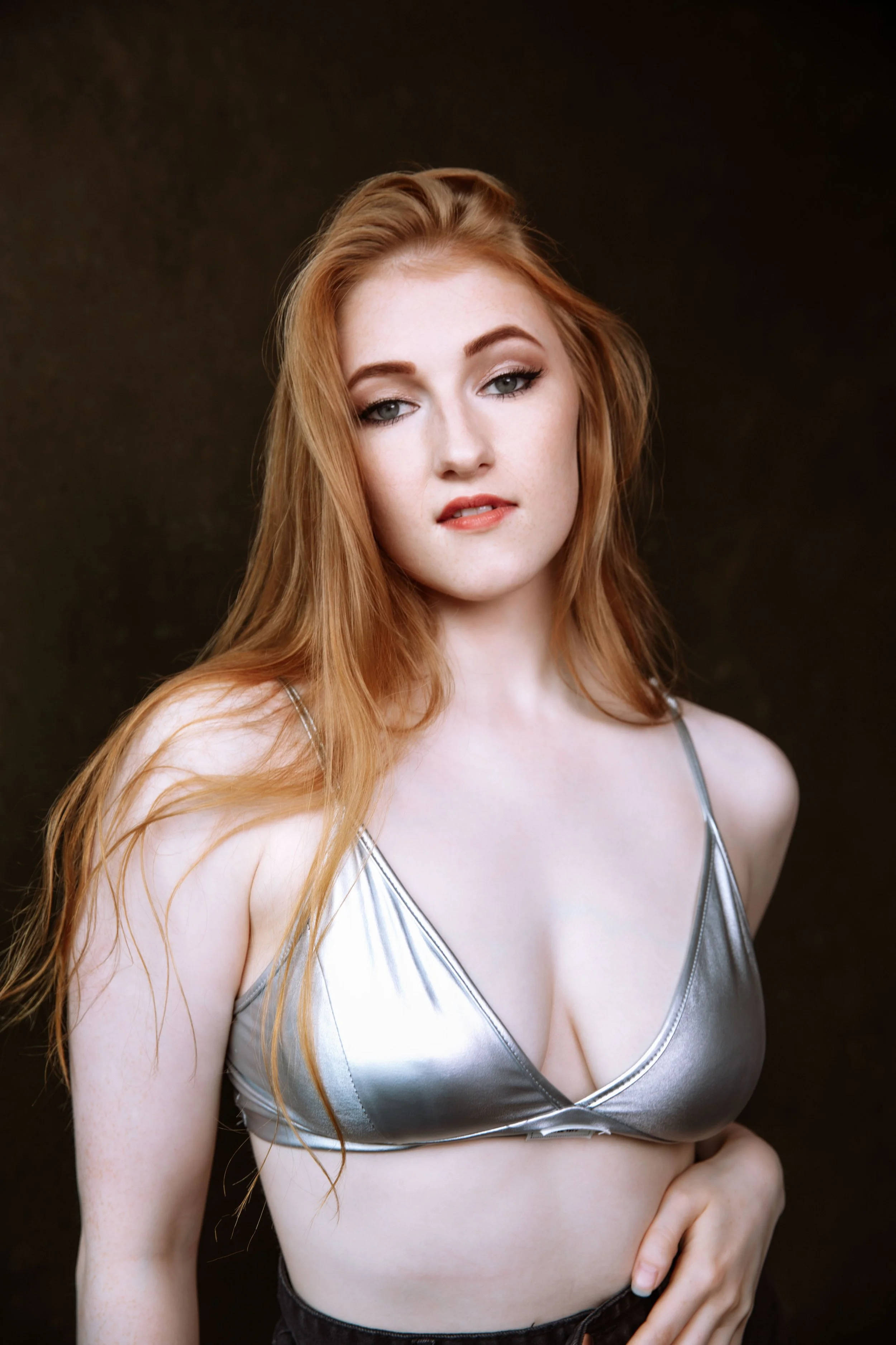 A young woman with long red hair and fair skin wearing a silver metallic top with thin straps, posing against a dark background.