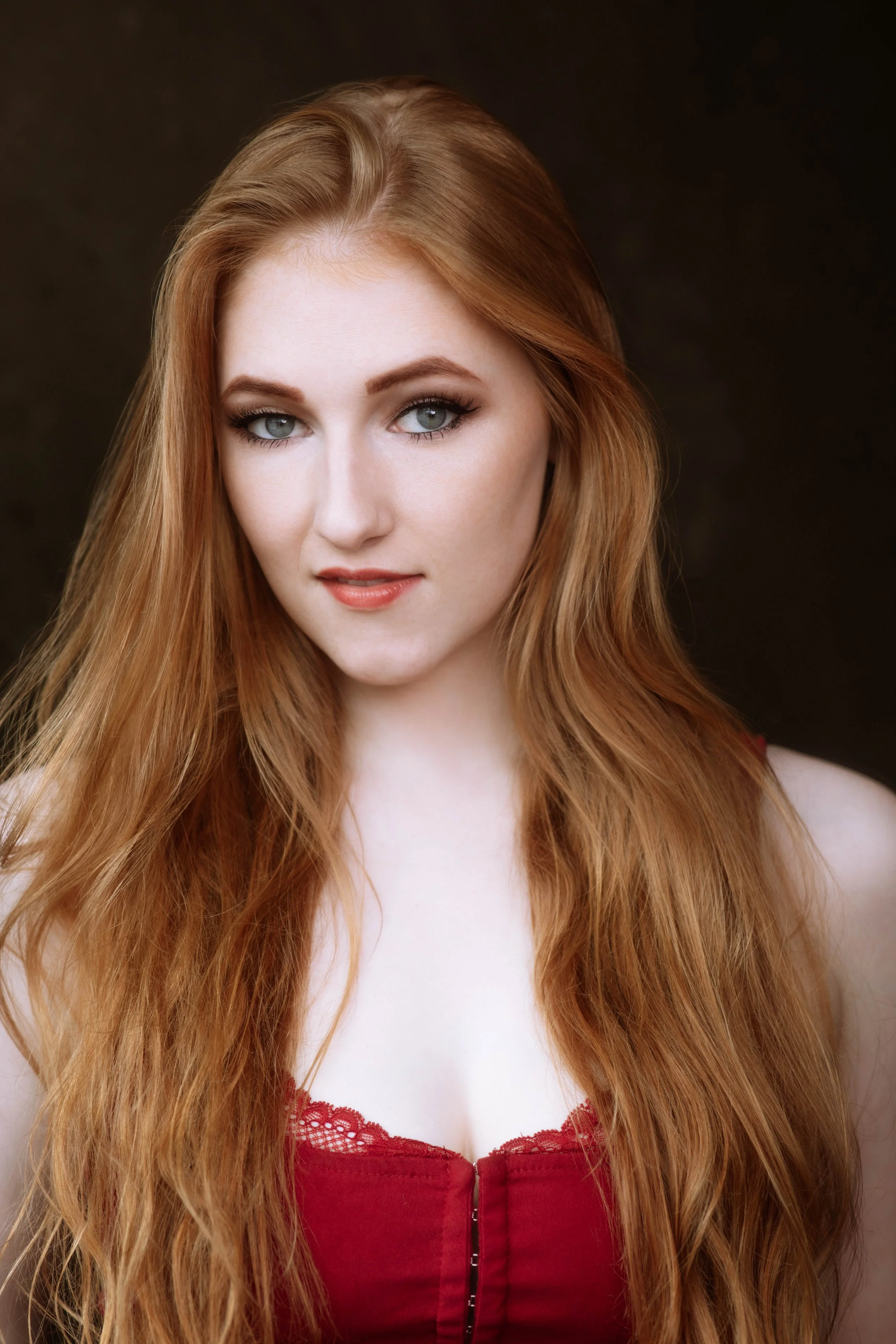 Portrait of a young woman with long, wavy red hair and blue eyes, wearing a red lace-trimmed top, against a dark background.