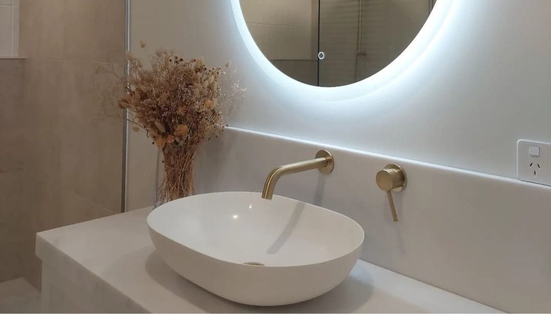 Modern bathroom counter with white oval basin, gold faucet, round backlit mirror, and dried flowers in a vase.