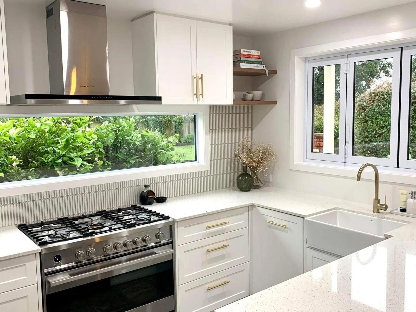 Modern kitchen with a stainless steel oven and range, white cabinets, white countertops, brass fixtures, and a large window showing greenery outside.