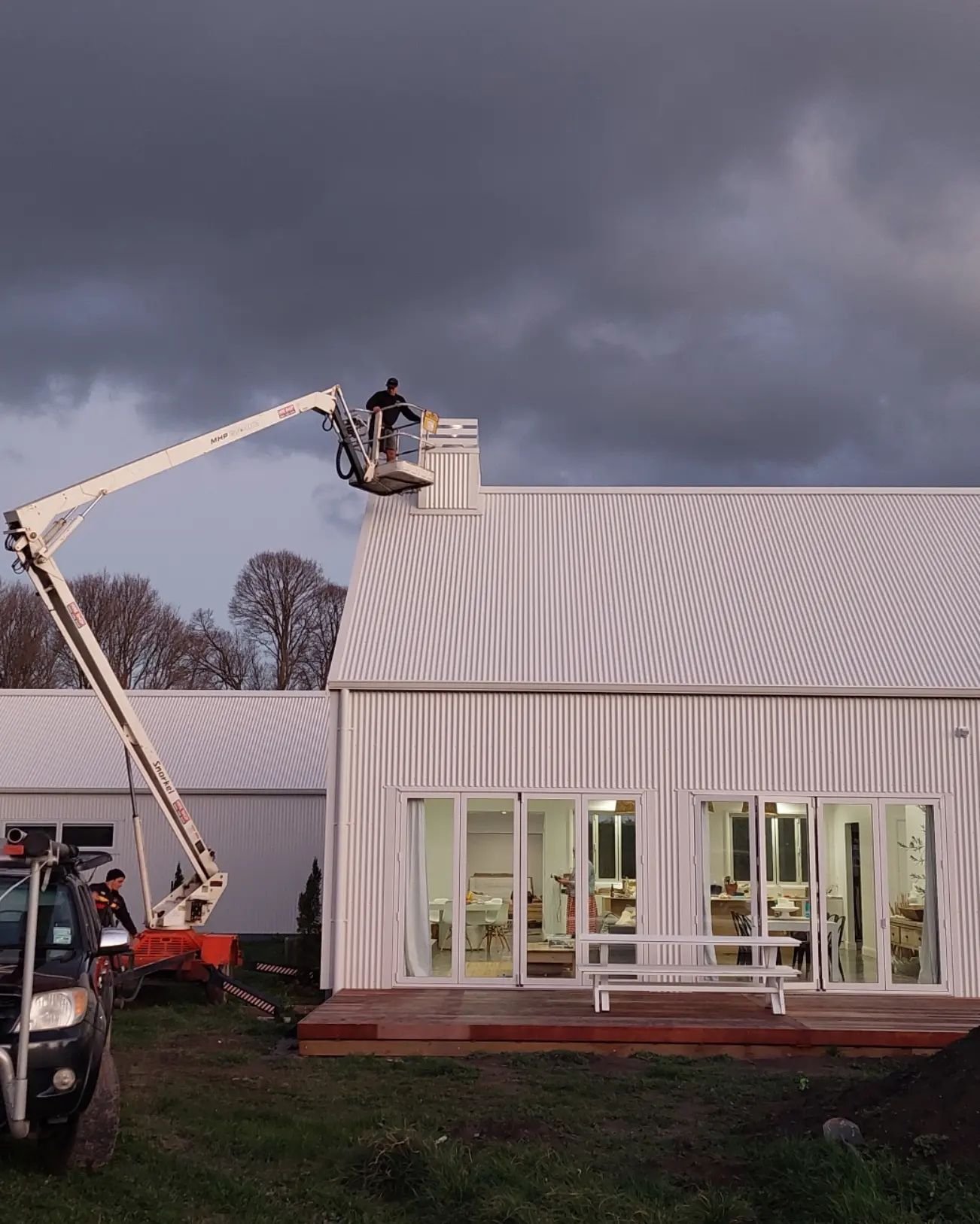 Person using a cherry picker to work on the roof of a modern white building on a cloudy day.