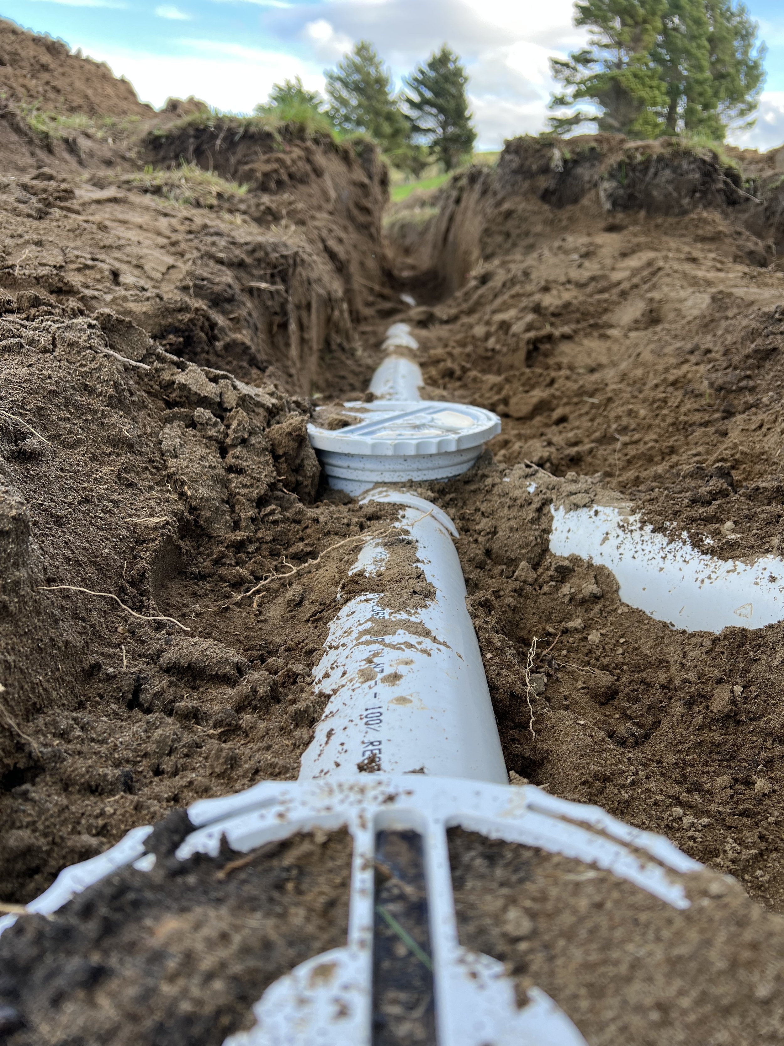 PVC drainage pipe in a newly dug trench with loose soil around it, set in an outdoor environment with trees in the background.