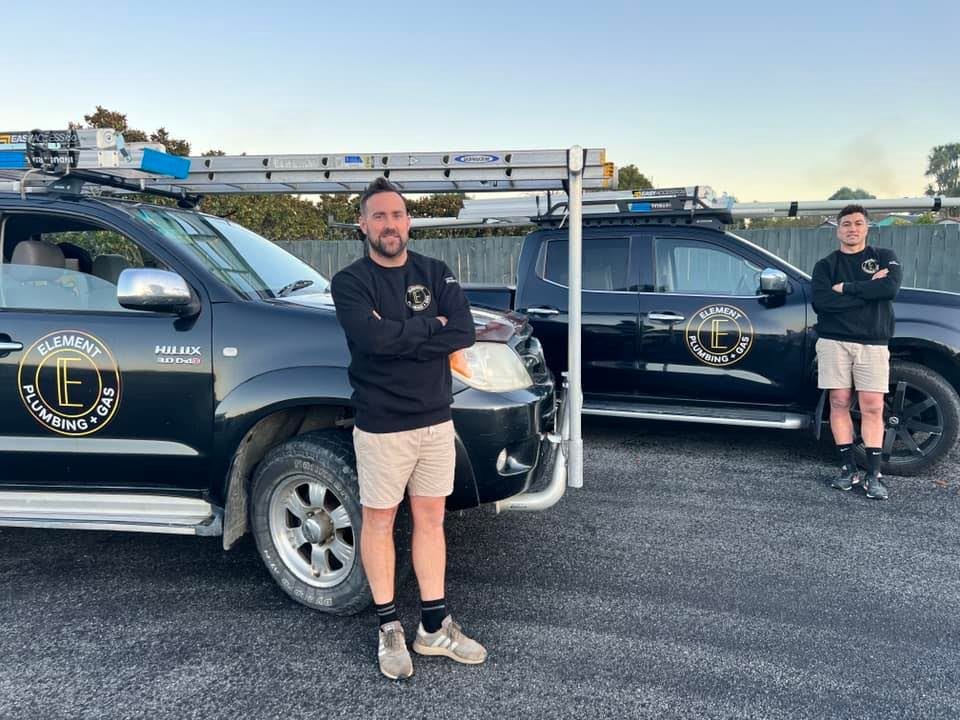 Two men standing in front of black pickup trucks with Element Plumbing and Gas branding, wearing work uniforms and standing on a paved area with ladders mounted on the trucks.