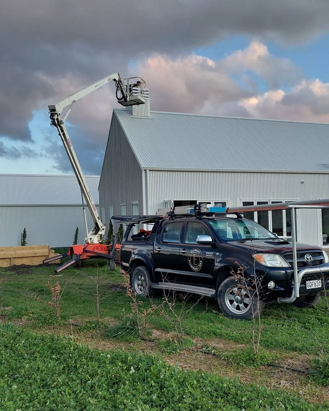 A black utility vehicle with a company logo parked on grass near a modern building. A boom lift with a person in the basket extends to the building's roof. The sky is partly cloudy.