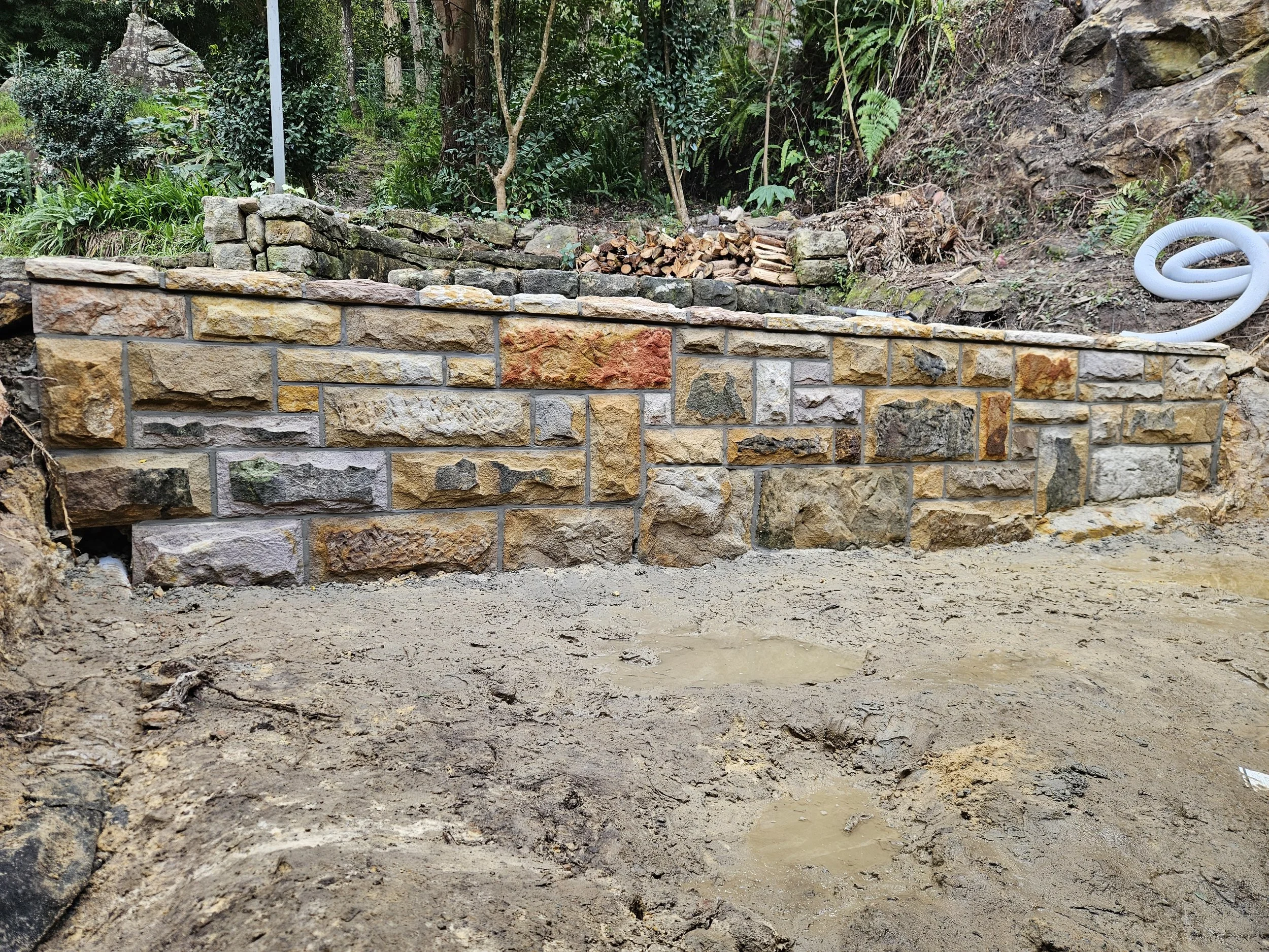 A stone and brick retaining wall built along a dirt and gravel area in a garden or backyard, with trees and shrubs in the background, a small pile of firewood above the wall, and a coiled white hose on the hillside to the right.