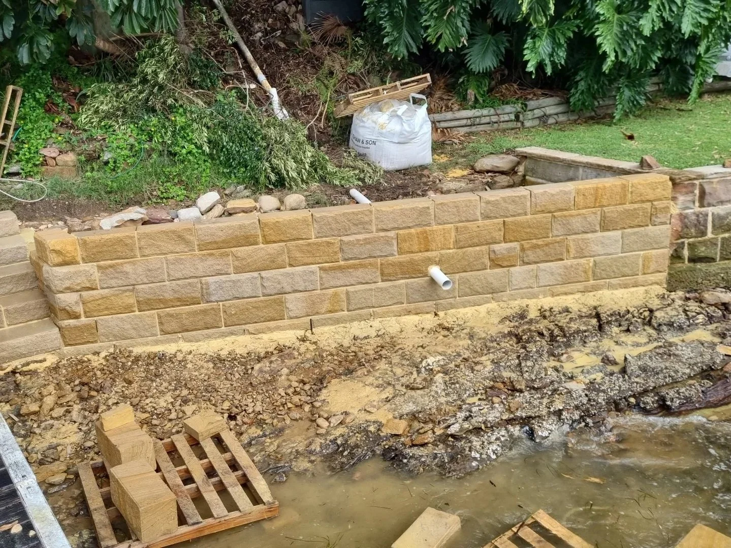 A brick retaining wall under construction along a waterfront, with earthy materials, a pipe sticking out, and building tools nearby.