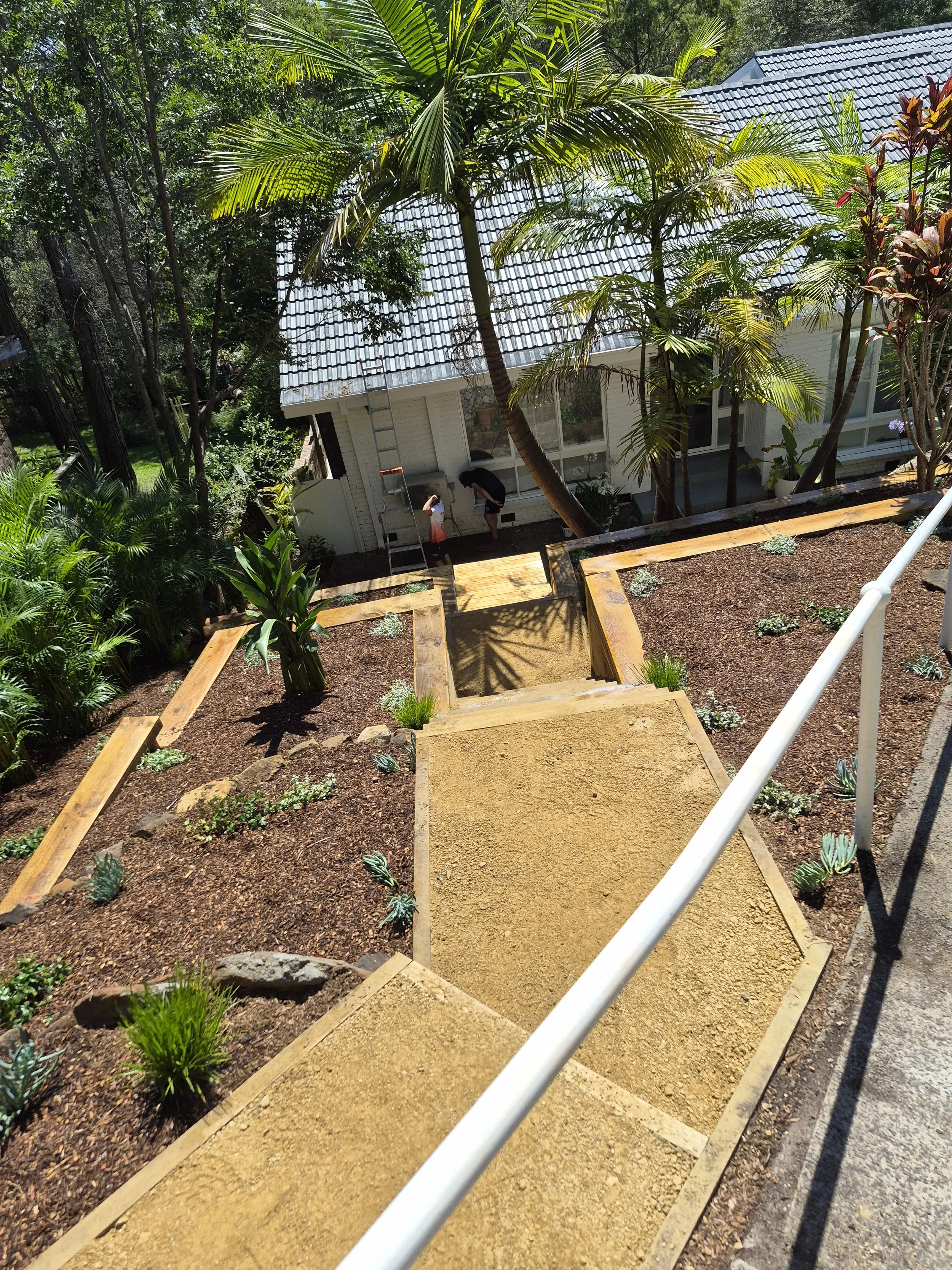 Person and child working on a garden pathway between two unfinished garden beds with wooden borders, tall palm trees, and a house with a metal roof in the background.