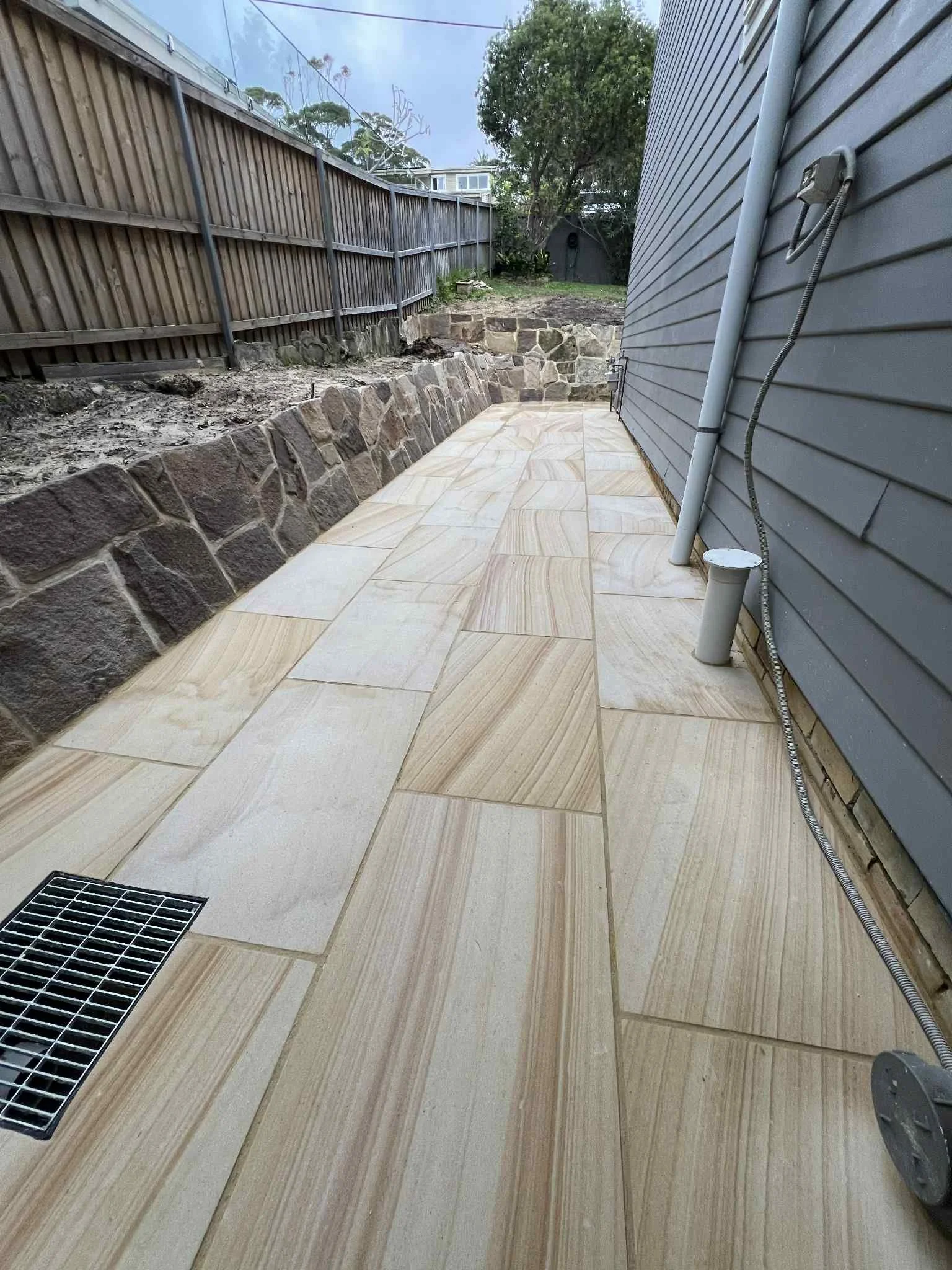 A backyard patio with a stone retaining wall along one side and beige tiled flooring. A gray house with exterior siding is on the right, with electrical wiring and tubing visible. A grate drain is on the patio, and a dirt area with a fence, trees, an