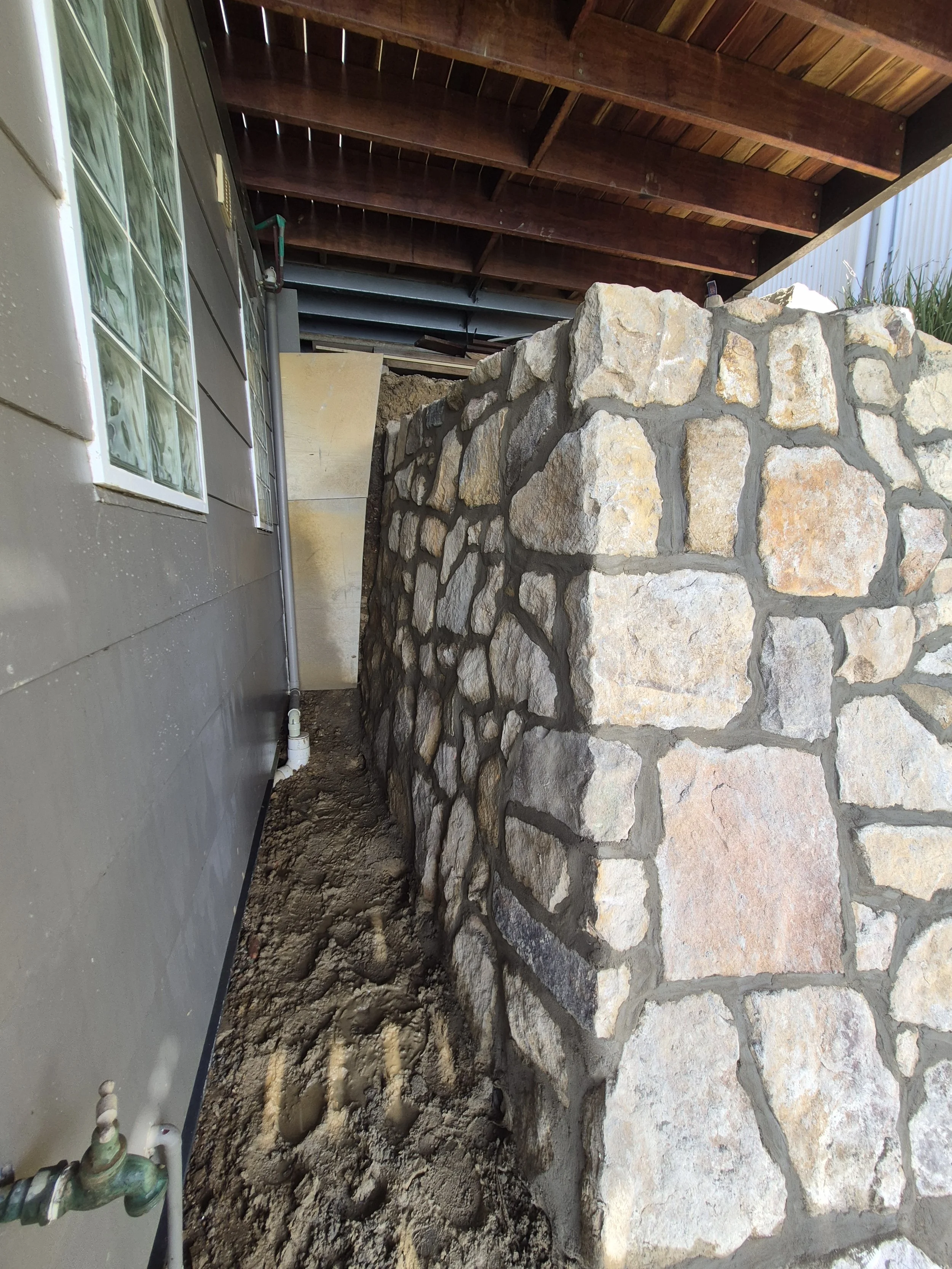 Image shows a stone retaining wall, a concrete foundation, a house with siding and windows, and a wooden roof extension.
