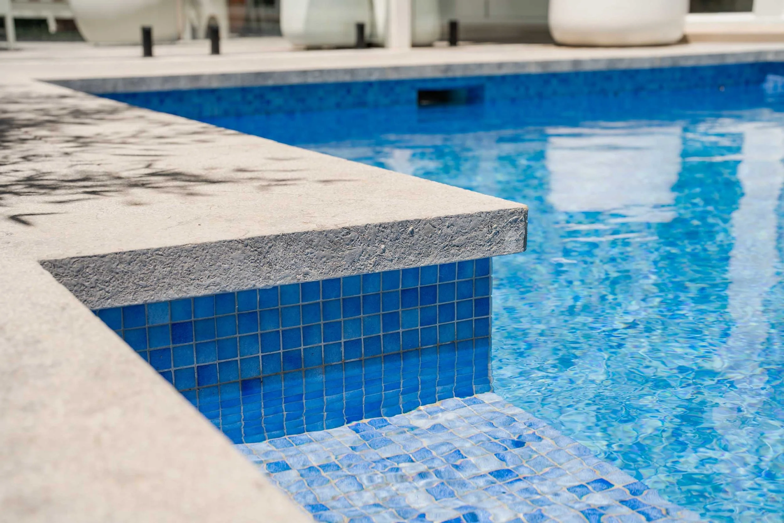 Close-up of the edge and steps of an outdoor swimming pool with blue tiles and clear water.