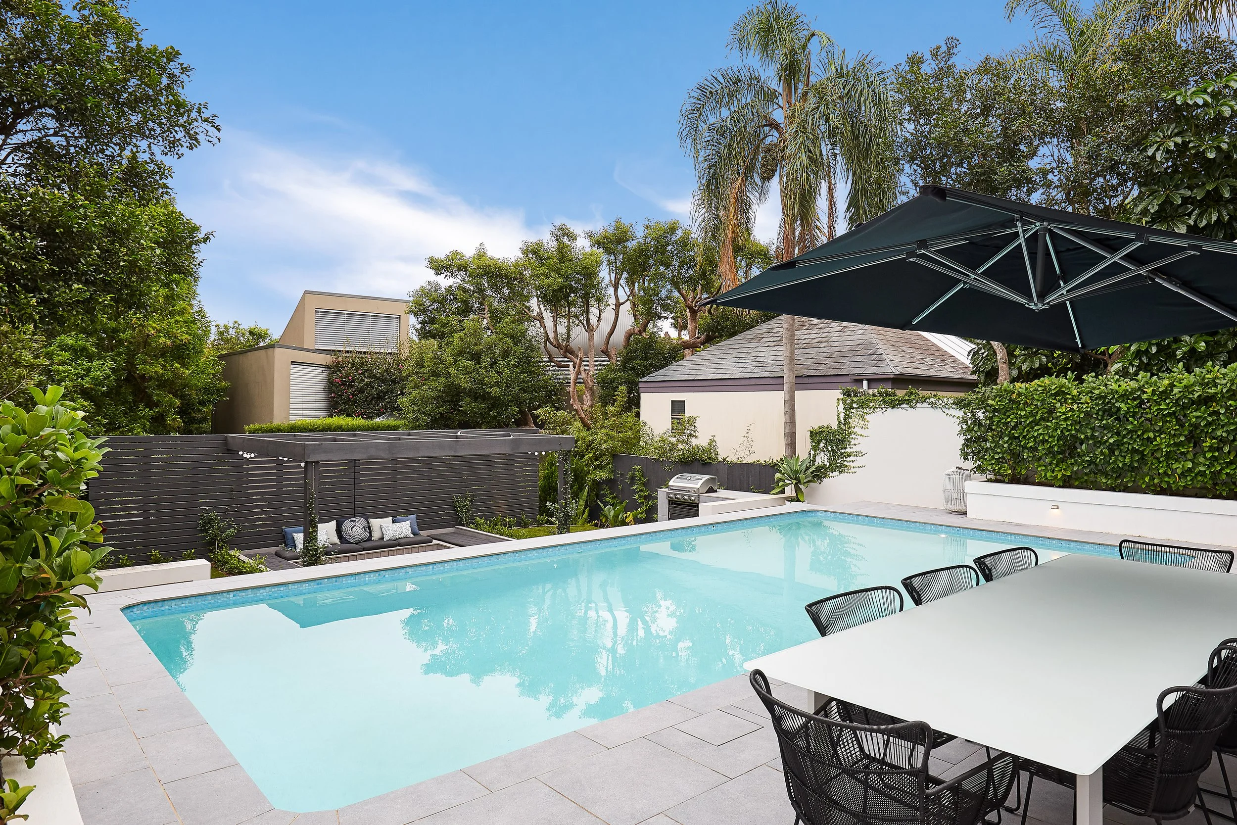 Garden with swimming pool, outdoor dining table with black chairs, large black patio umbrella, lush green trees and bushes, and modern buildings in the background under a blue sky.