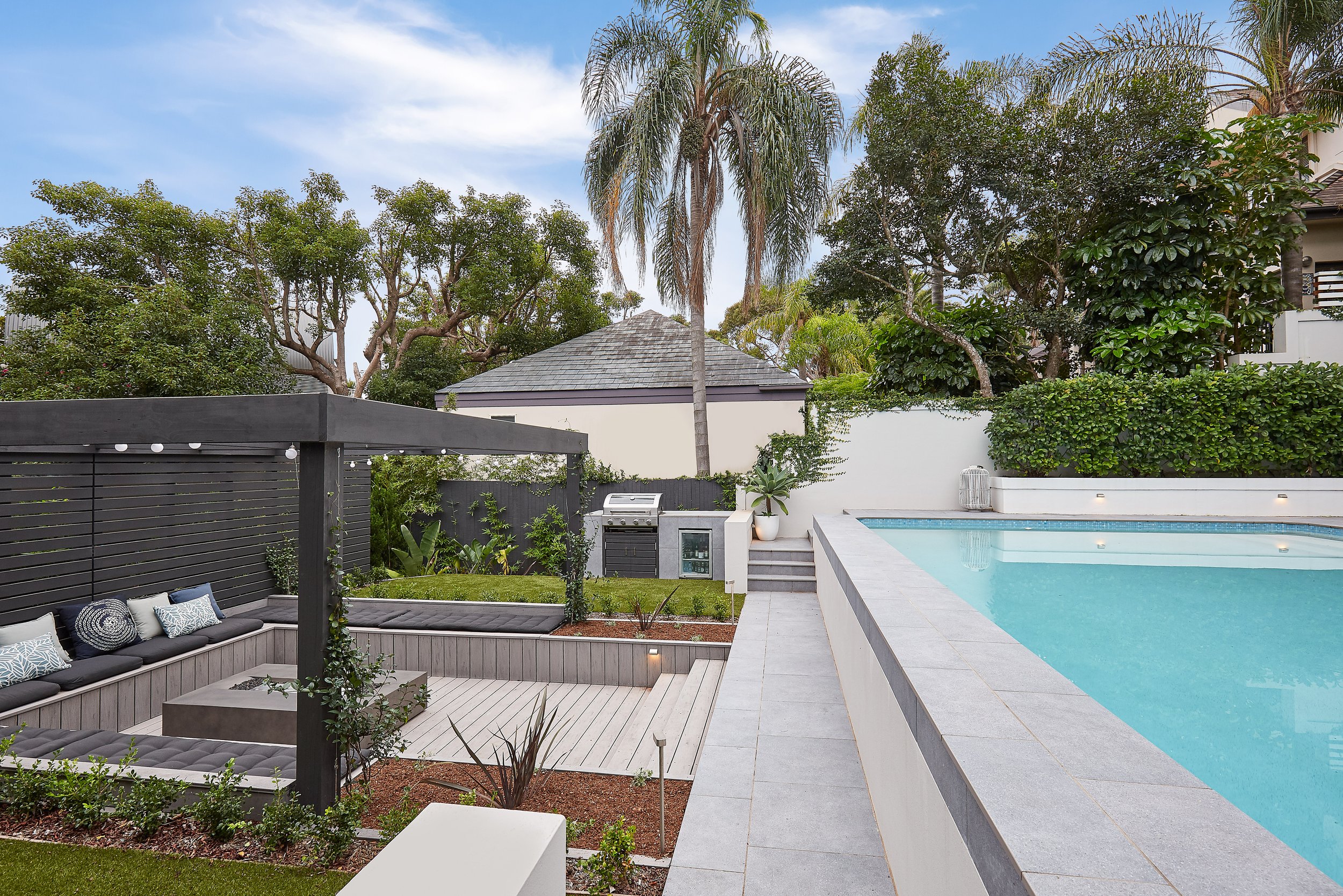 Modern garden with a swimming pool, outdoor seating area with cushions, and lush greenery, including trees and shrubs and a firepit.