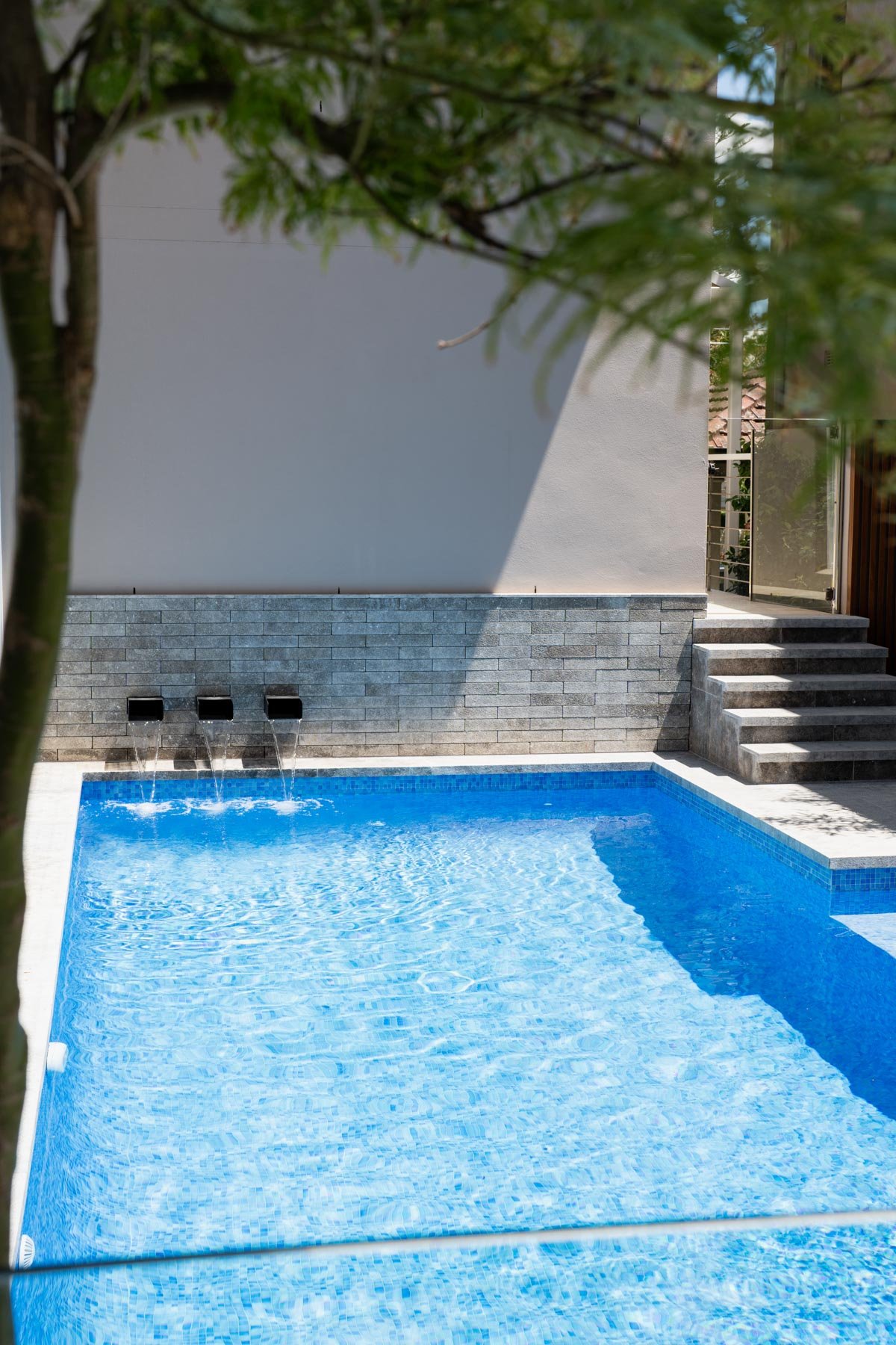 Empty backyard swimming pool with clear blue water, gray stone wall with three water spouts, gray concrete stairs leading up to a door, partially obscured by tree branches.